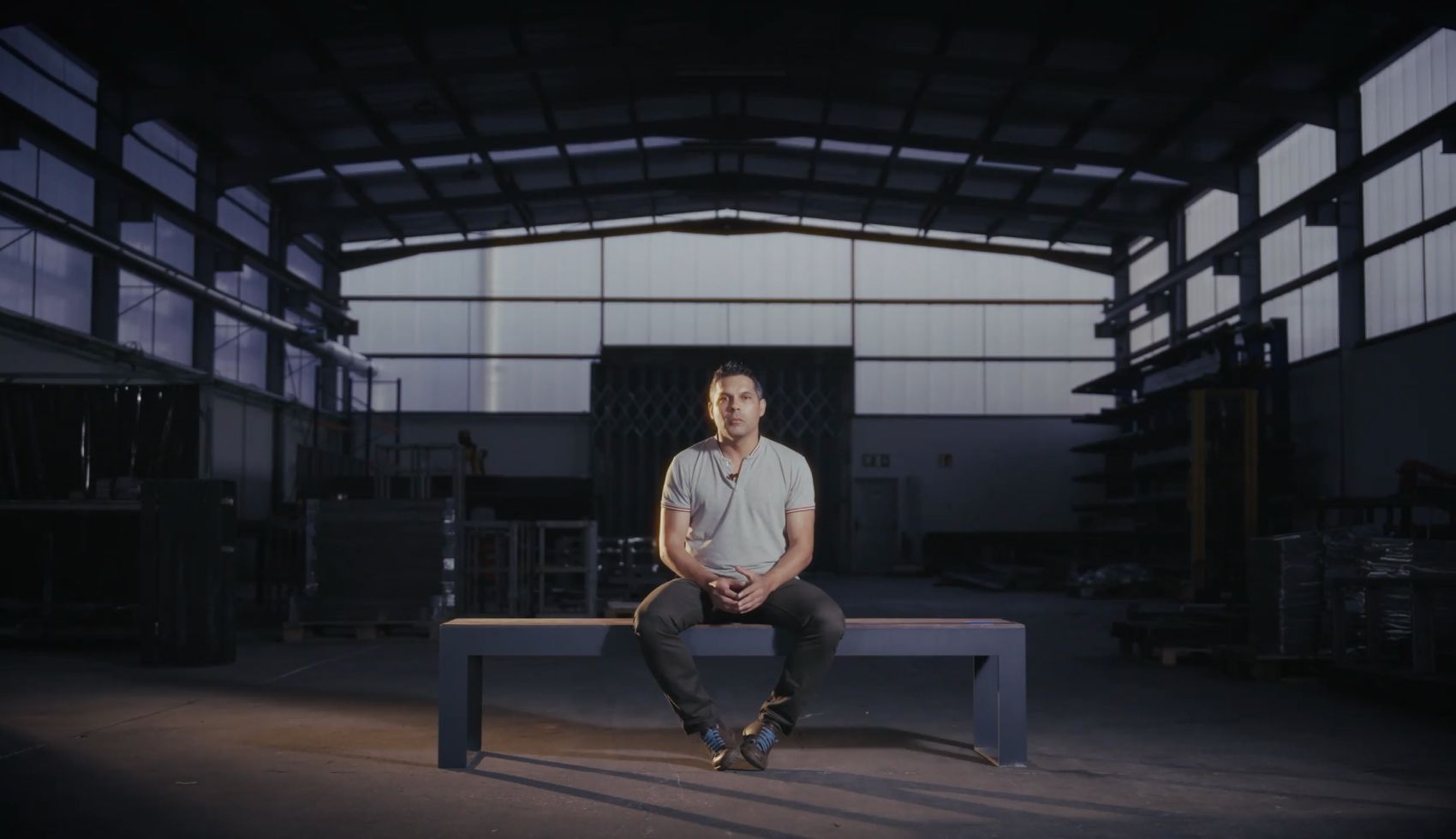 Designer de produto Nelson António sentado no banco sem costas Frame da Floema Urban com o pavilhão de serralharia da unidade de produção Floema como fundo.